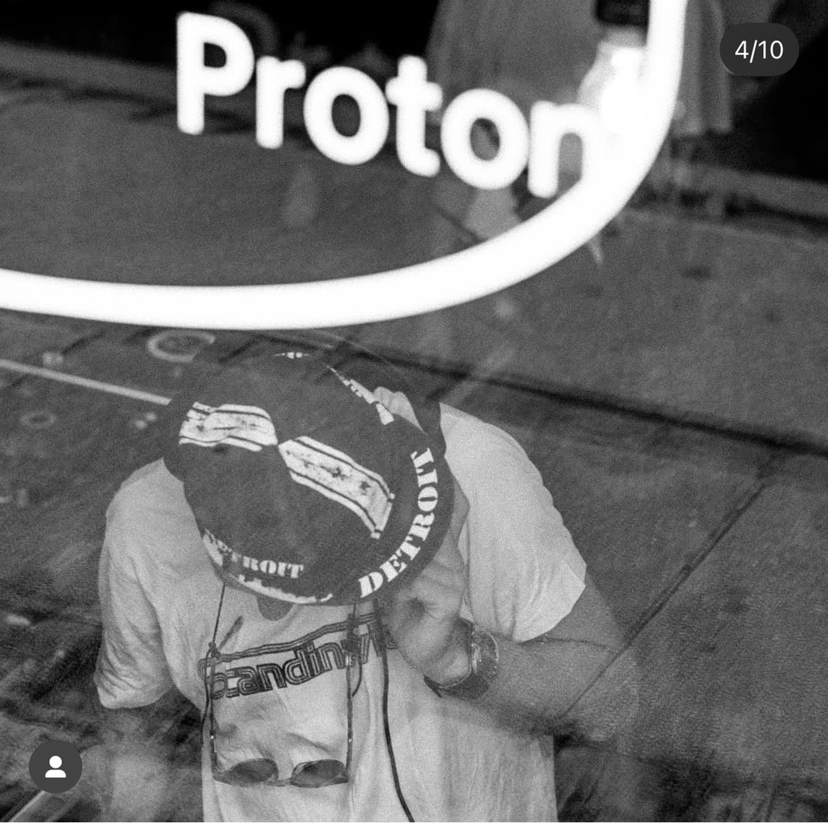 A person in a Detroit bucket hat, seen through a glowing Proton neon sign